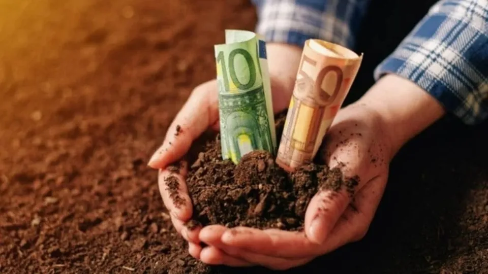 Două mâini țin un pământ întins, în care sunt introduse două bancnote rulate, una verde și una portocalie.