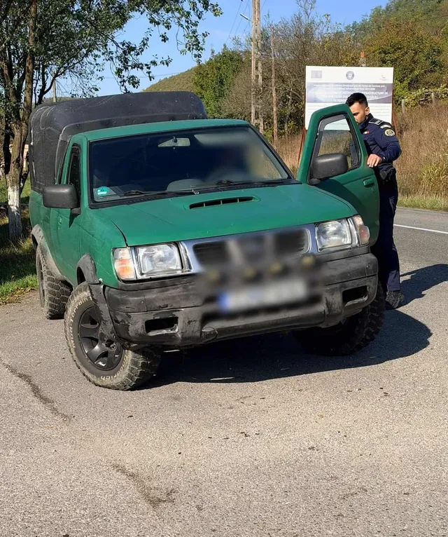 Un bărbat în uniformă întunecată stă lângă portiera deschisă a unui pickup verde parcat pe marginea unui drum asfaltat.