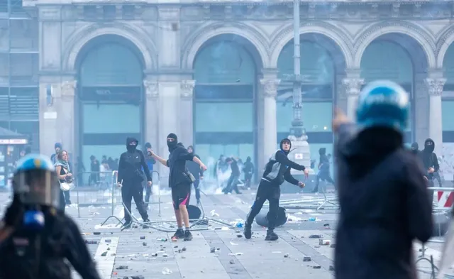 O mulțume de persoane îmbrăcate în negru, unele cu cască, se află pe o piață cu arce de fundal, aruncând obiecte într-un mediu cu praf și detritus.