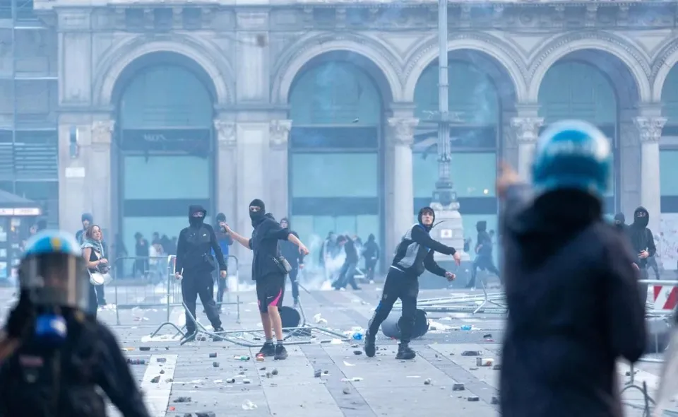 O mulțume de persoane îmbrăcate în negru, unele cu cască, se află pe o piață cu arce de fundal, aruncând obiecte într-un mediu cu praf și detritus.