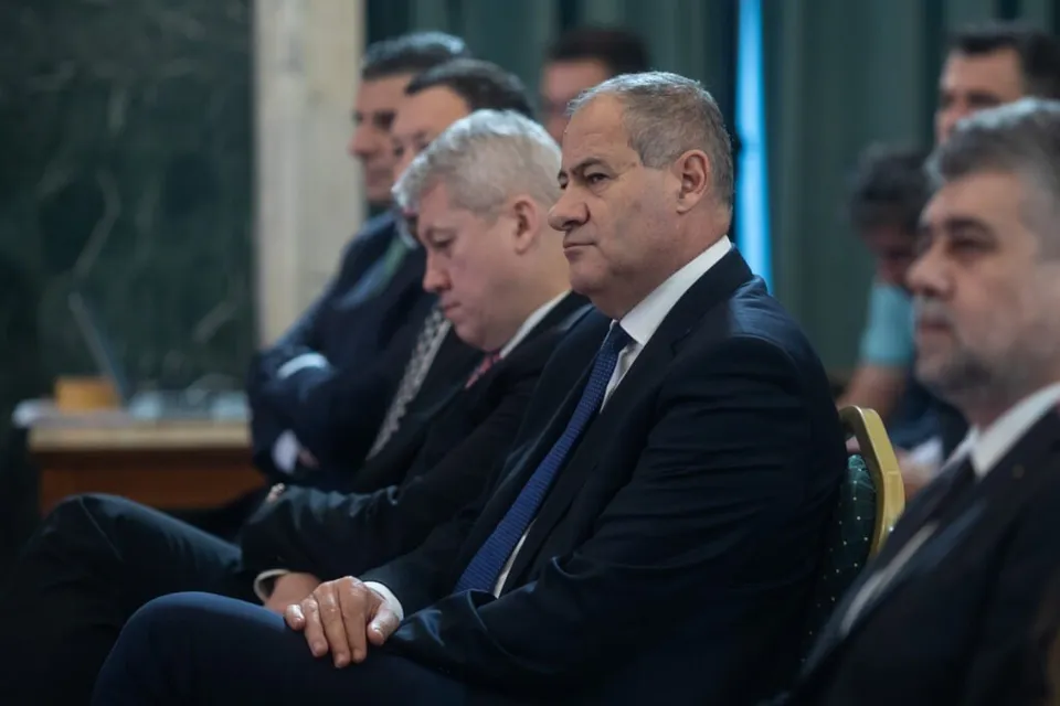 Cinci bărbați în costume și cravate stau așezați pe scaune într-o încăpere interioară.
