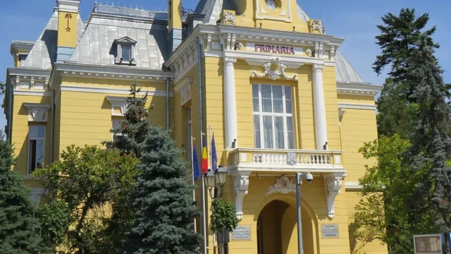 O clădire galbenă cu elemente arhitecturale albe, coloane și ferestre mari, înconjurată de copaci verzi sub un cer senin.