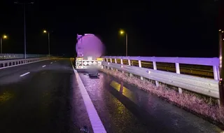 O autocisternă și un autoturism sunt parcate pe marginea unei autostrăzi noapte, lângă un guardrail, sub lumina stâlpilor de iluminat public.