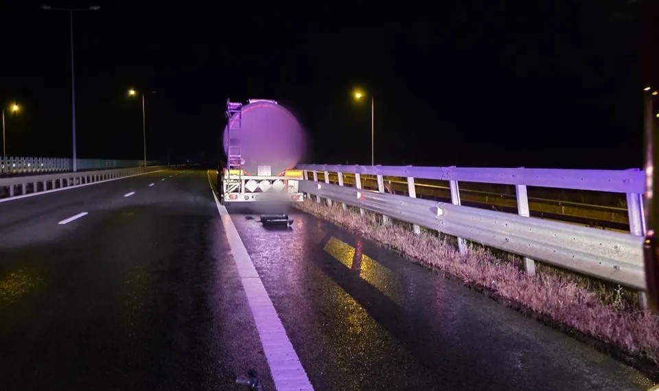 O autocisternă și un autoturism sunt parcate pe marginea unei autostrăzi noapte, lângă un guardrail, sub lumina stâlpilor de iluminat public.