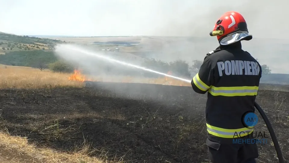 Un pompier cu echipament de protecție și cască roșie pulverizează un jet de apă pe un teren de vegetație arsă sub un cer înnorat.