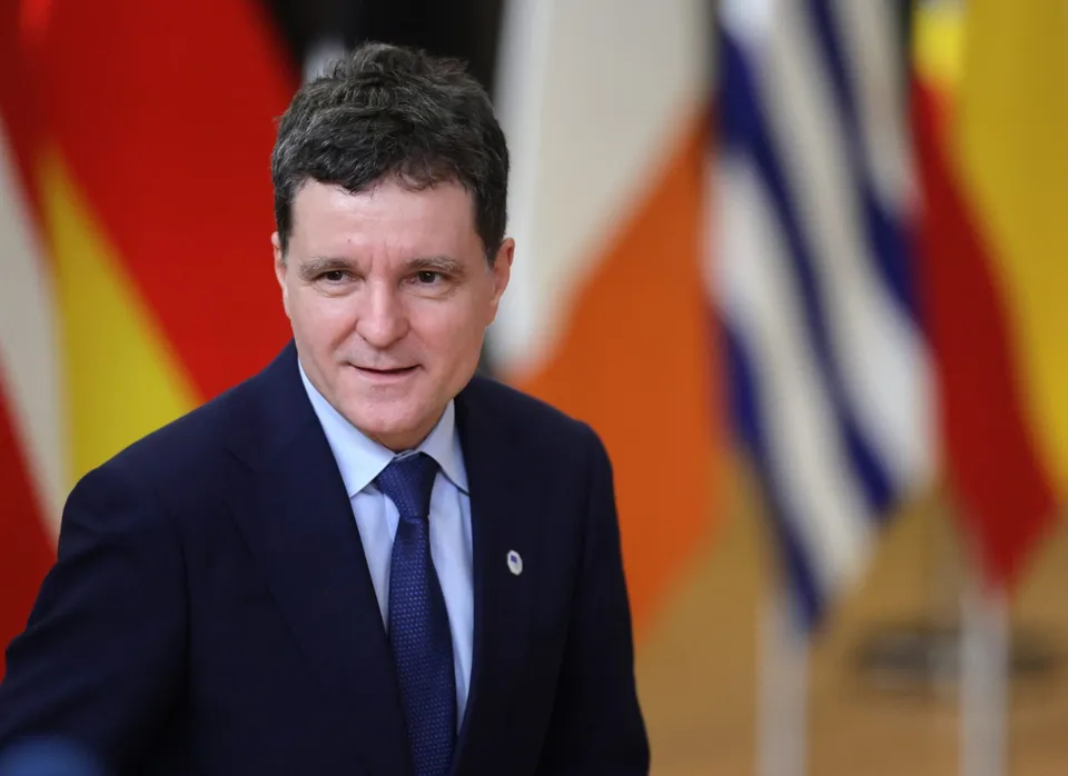 Un bărbat cu părul scurt și închis la culoare poartă un costum închis, cămașă albastră și cravată, stând în fața unor steaguri colorate.