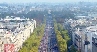 O mulțime de oameni este adunată pe o bulevardă lungă, încadrată de copaci și clădiri înalte, sub un cer senin.