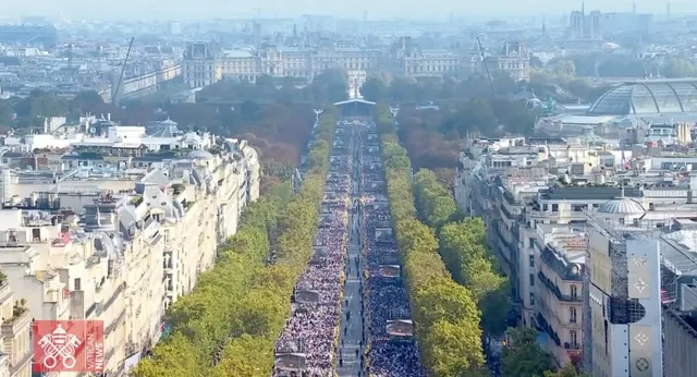 O mulțime de oameni este adunată pe o bulevardă lungă, încadrată de copaci și clădiri înalte, sub un cer senin.
