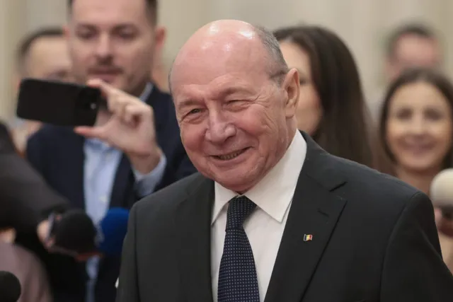 Un bărbat vârstnic cu părul rare, purtând costum negru, cămașă albă și cravată neagră, zâmbește în timp ce este înconjurat de persoane în plan secund.