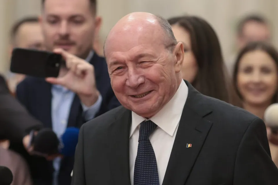 Un bărbat vârstnic cu părul rare, purtând costum negru, cămașă albă și cravată neagră, zâmbește în timp ce este înconjurat de persoane în plan secund.