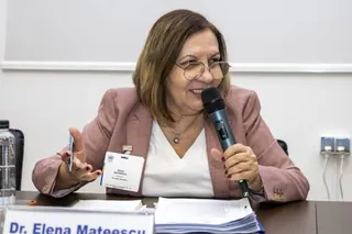 O femeie cu ochelari și sacou roz vorbește într-un microfon, stând la o masă cu documente și un badge pe piept.