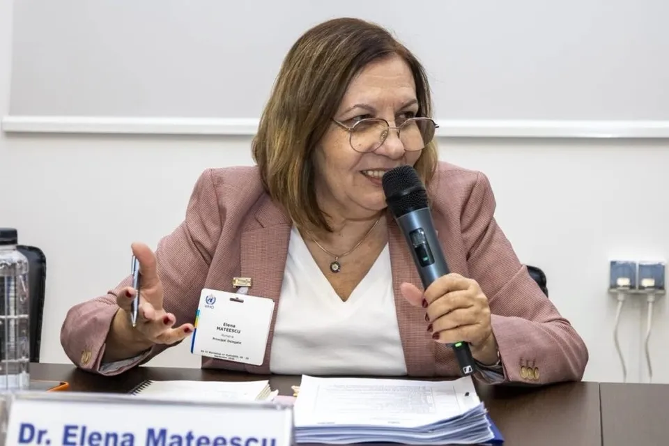 O femeie cu ochelari și sacou roz vorbește într-un microfon, stând la o masă cu documente și un badge pe piept.