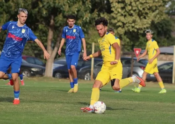 Câțiva jucători de fotbal în echipamente albastre și galbene joacă pe un teren de iarbă verde sub lumina soarelui.