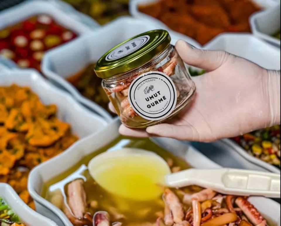 O mână cu mănușă albă ține un borcan de sticlă cu capac metalic deasupra unor tăvi cu diverse alimente preparate.