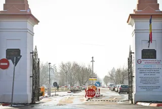 O poartă metalică neagră se află între doi piloni albi, deschizând drumul spre o zonă cu copaci și vehicule parcate în depărtare.
