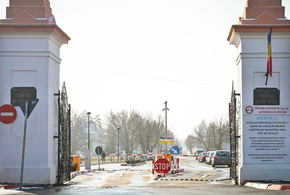 O poartă metalică neagră se află între doi piloni albi, deschizând drumul spre o zonă cu copaci și vehicule parcate în depărtare.