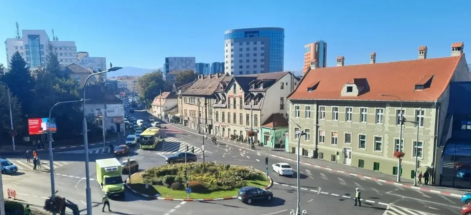 O vedere de sus a unei intersecții urbane cu mașini, un autobuz galben, un rond de sens giratoriu și clădiri pe fundal sub un cer senin.