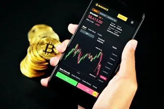 O mână ține un smartphone care afișează un grafic de tranzacționare criptomonede lângă un grup de monede aurii pe un fundal negru.