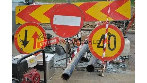O barieră de lucru cu bandă galie și roșie, trei semnale de circulație (interdicție, sens interzis, limită de viteză 40) și țevi de metal pe un șantier.