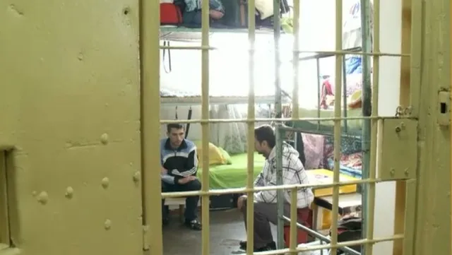 Doi bărbați stau așezați într-o cameră cu paturi prin grilajul unei uși metalice deschise.