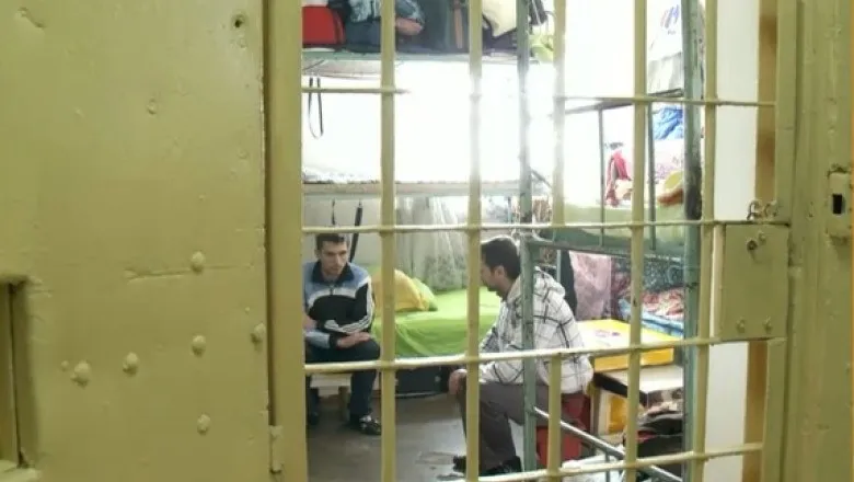 Doi bărbați stau așezați într-o cameră cu paturi prin grilajul unei uși metalice deschise.