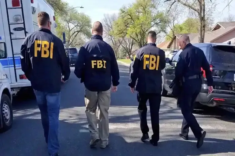 Patru bărbați cu inscripții FBI pe haine merg pe un drum lângă un vehicul alb și o mașină neagră.