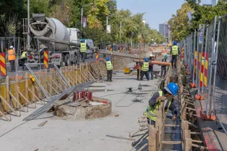 O echipă de muncitori cu căști și veste reflectorizante lucrează pe un șantier de construcții lângă un camion cu betonieră.