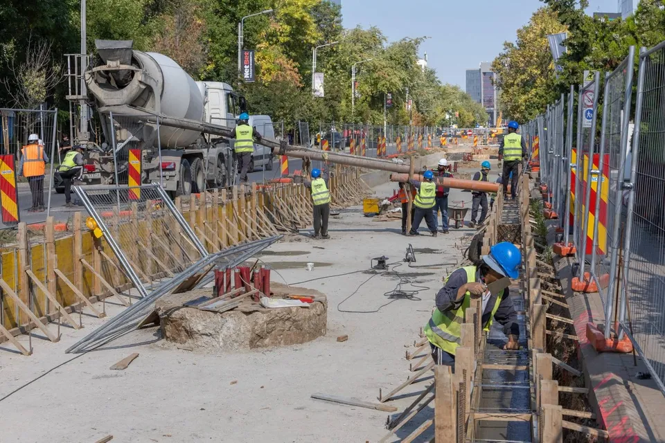 O echipă de muncitori cu căști și veste reflectorizante lucrează pe un șantier de construcții lângă un camion cu betonieră.