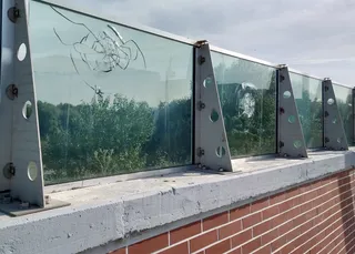 Panouri de sticlă transparente cu crăpături sunt montate pe suporturi metalice gri deasupra unui zid de cărămidă roșie.