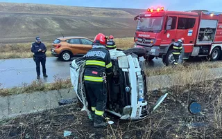 Un autoturism alb este răsturnat pe o parte în vegetație, lângă un camion de pompieri roșu și un autoturism portocaliu pe un drum asfaltat.