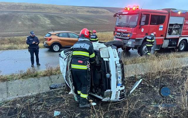 Un autoturism alb este răsturnat pe o parte în vegetație, lângă un camion de pompieri roșu și un autoturism portocaliu pe un drum asfaltat.