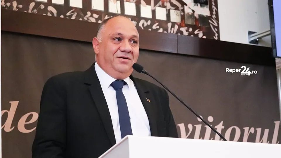 Un bărbat cu costum negru, cămașă albă și cravată albastră stă în spatele unui podium lângă un microfon.