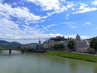 Un râu curge pe lângă un oraș cu clădiri albe, un pod și un castel situat pe un deal sub un cer cu nori albi.