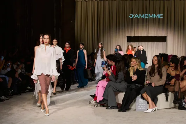 O femeie merge pe o pistă de catwalk purtând o bluză albă voluminoasă și o fustă roz, în timp ce spectatori stau pe scaune în lateral.