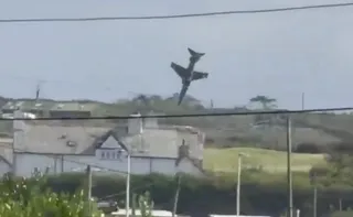 Un avion de culoare închisă zboară vertical în cer deasupra unui peisaj cu dealuri și clădiri albe.