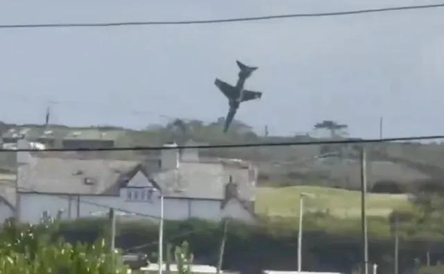 Un avion de culoare închisă zboară vertical în cer deasupra unui peisaj cu dealuri și clădiri albe.