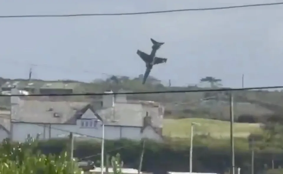 Un avion de culoare închisă zboară vertical în cer deasupra unui peisaj cu dealuri și clădiri albe.