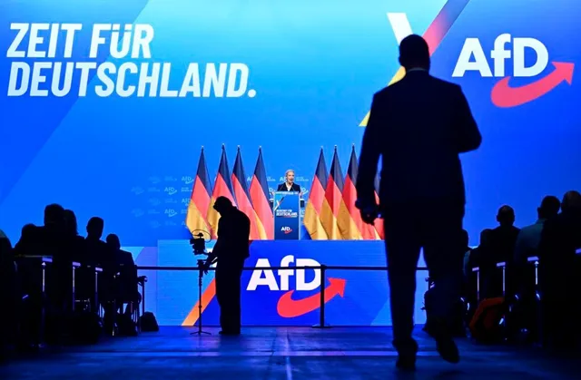 Un bărbat în costum merge spre un podium pe care sunt așezate câteva steaguri germane, în fața unui panou albastru cu text și un logo.