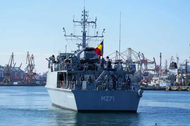 Un vas gri cu numărul M271 navighează pe apă, având un steag tricolor și macarale de port în fundal sub un cer senin.