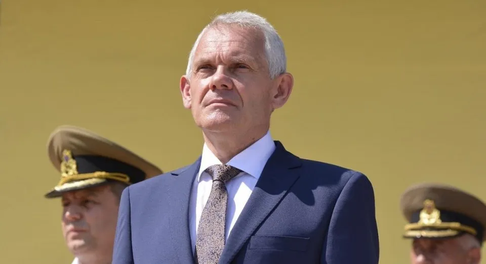 Un bărbat cu părul scurt și alb, purtând un costum albastru, cămașă albă și cravată gri, privește în sus în fața unui fundal galben.