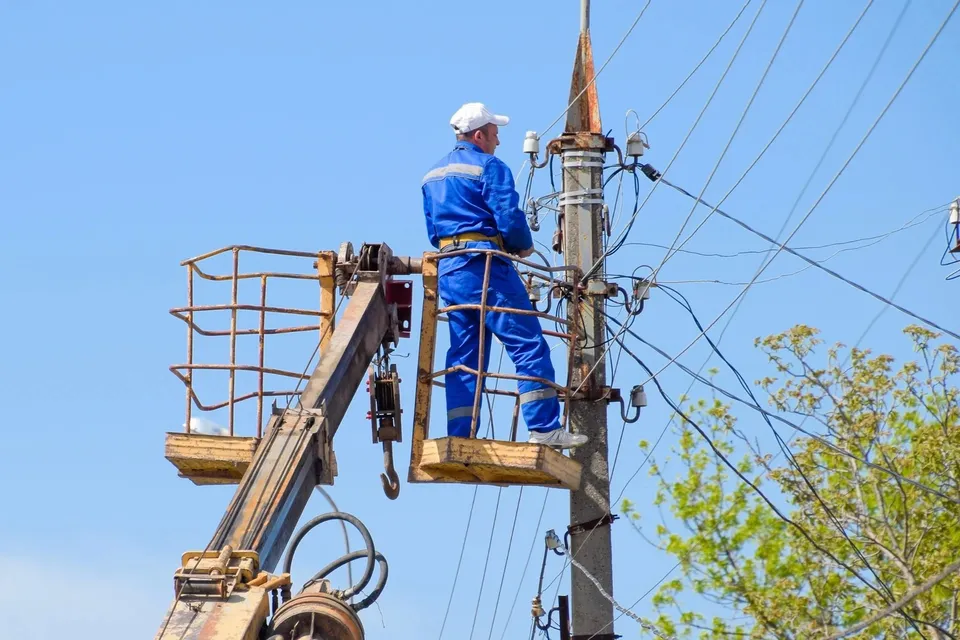 Un bărbat cu cască și costum albastru stă într-un coș de lucru suspendat de un utilaj lângă un stâlp electric sub un cer senin.