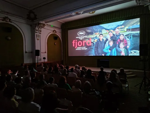 Oamenii stau în întuneric într-o sală de cinema, privind un ecran mare pe care este proiectată o imagine cu un grup de persoane într-un peisaj montan.