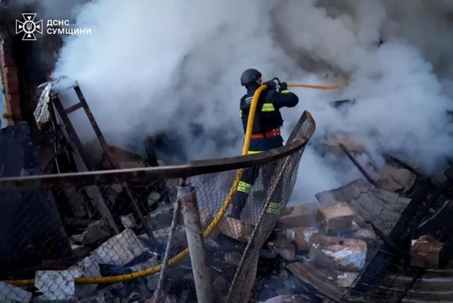 Un pompier cu echipament de protecție și cască folosește un furtun galben pentru a stinge un incendiu printre dărâmături și fum dens.
