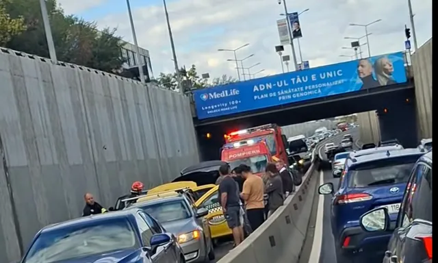Autoturisme și un camion de pompieri sunt oprite într-o coadă de trafic pe un drum sub un pod cu un afiș publicitar deasupra.