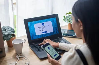 O persoană ține un smartphone lângă un laptop deschis pe o masă de lemn lângă o cană albă și o plantă.