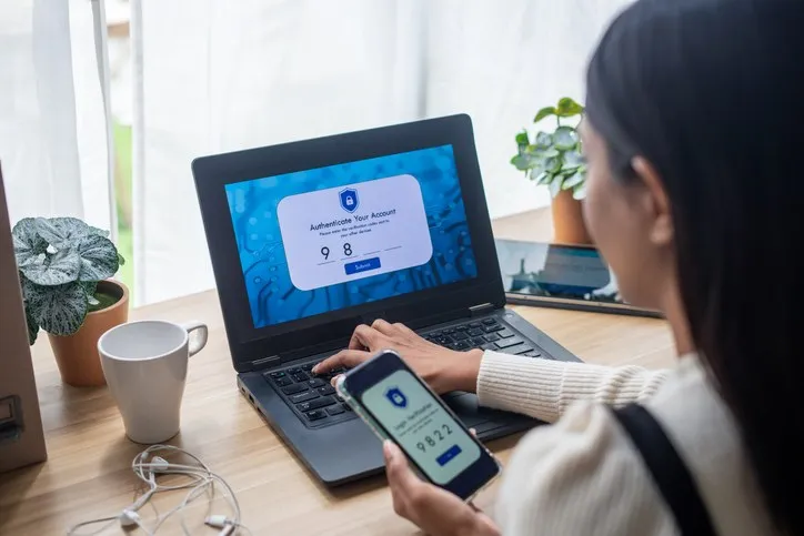 O persoană ține un smartphone lângă un laptop deschis pe o masă de lemn lângă o cană albă și o plantă.