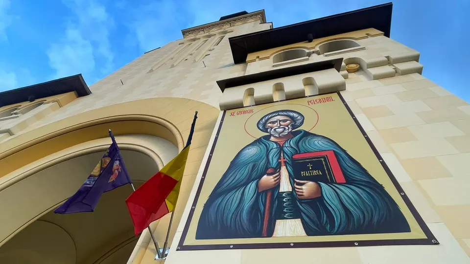 O pictură murală a unui bărbat cu barbă, purtând o robă albastră și ținând un obiect dreptunghiular roșu, este afișată pe fațada unei clădiri.