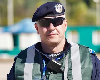 Un bărbat poartă beretă neagră, ochelari de soare și o vestă reflectorizantă peste o uniformă cu inscripția Jandarmeria.