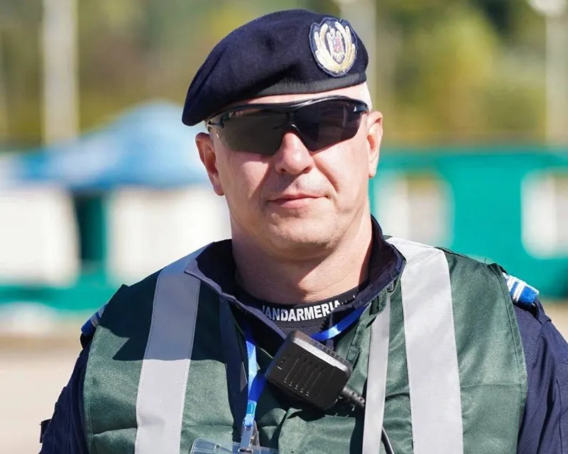 Un bărbat poartă beretă neagră, ochelari de soare și o vestă reflectorizantă peste o uniformă cu inscripția Jandarmeria.