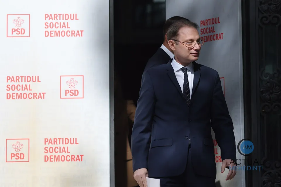 Un bărbat cu ochelari și costum închis la culoare pășește printr-o ușă lângă un panou alb cu logoul PSD repetat de mai multe ori.